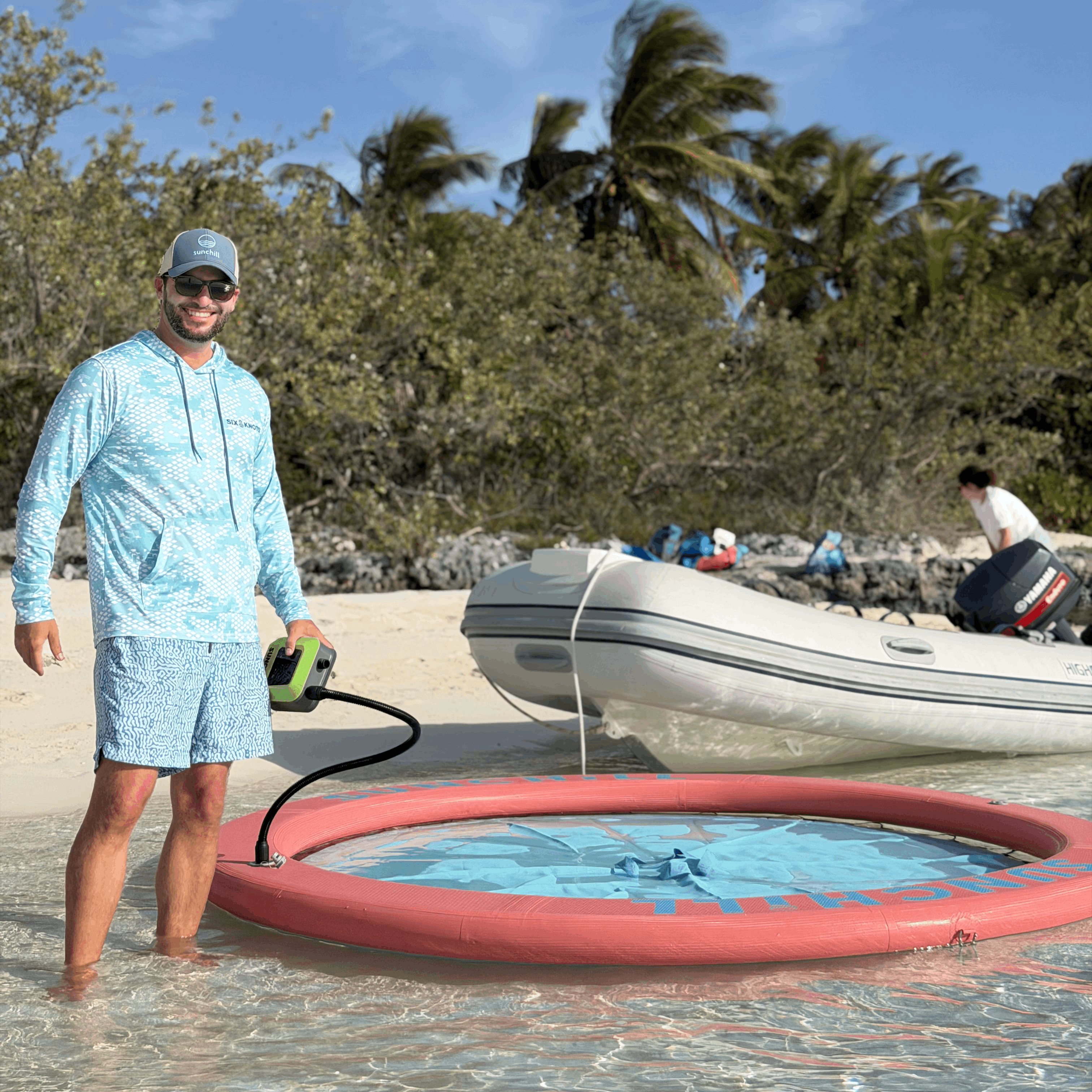 Man operating a portable electric pump on a beach with a boat and palm trees in the background